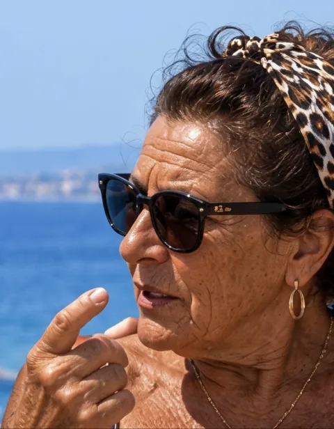 A Mameta in a leopard-print headband and sunglasses by the sea, one finger raised mid-sentence